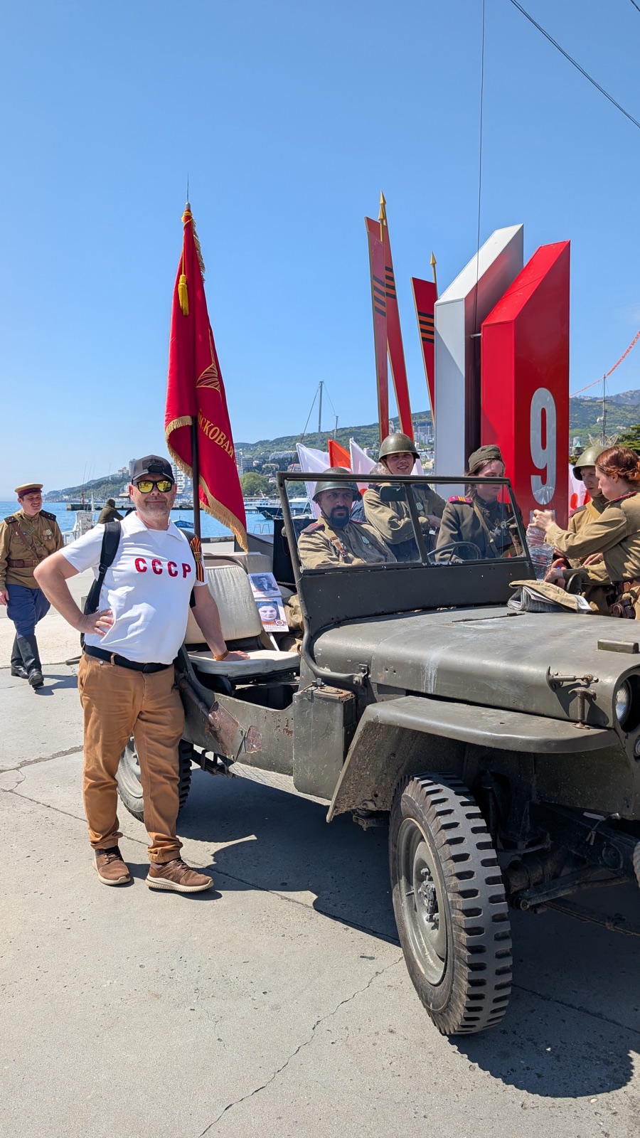 Reiseteilnehmer neben einem historischen Geländewagen vor der Bühne zum 9. Mai