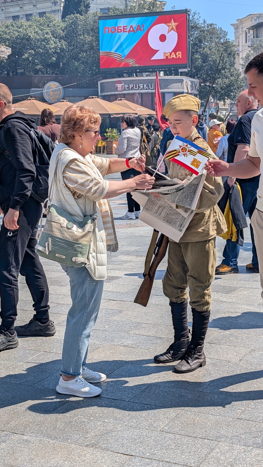 Junge in historischer Uniform verteilt Zeitungen an eine Passantin
